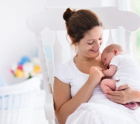 Young mother holding her newborn child. Mom nursing baby. Woman and new born boy relax in a white bedroom with rocking chair and blue crib. Mom relaxing to represent Recovering After C Section Surgery