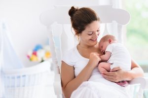 Young mother holding her newborn child. Mom nursing baby. Woman and new born boy relax in a white bedroom with rocking chair and blue crib. Mom relaxing to represent Recovering After C Section Surgery