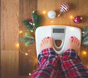 Top view of female feet in winter pajamas on digital scales or weight scale on wooden background surrounded with Christmas lights and decoration to represent the battle of the holiday bulge and weight gain