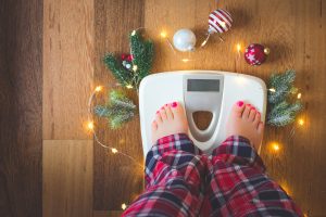 Top view of female feet in winter pajamas on digital scales or weight scale on wooden background surrounded with Christmas lights and decoration to represent the battle of the holiday bulge and weight gain