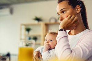 Young woman suffering from postpartum depression and thinking of something while being with her baby at home.