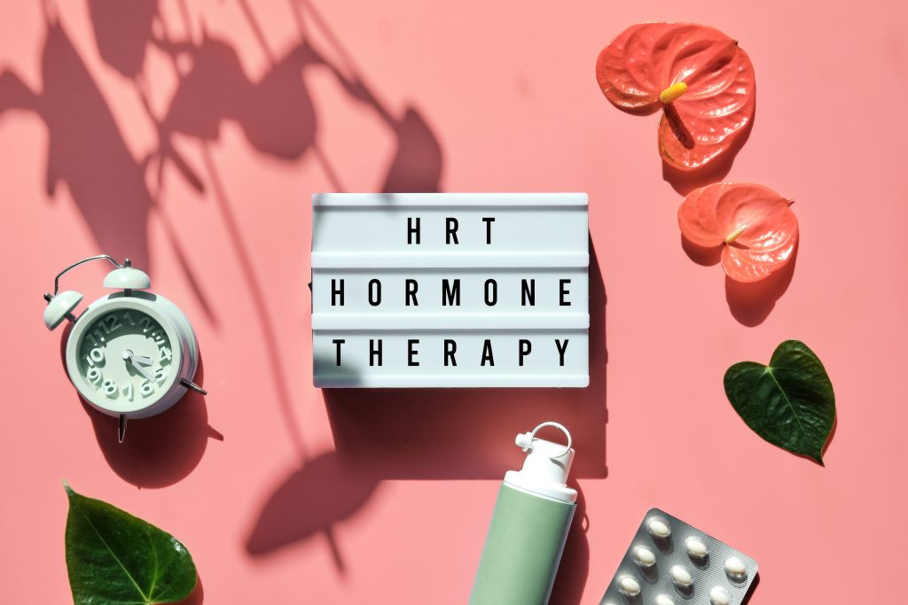 Text HRT Replacement Therapy on light box. Menopause, hormone therapy concept. Oestrogen replacement therapy awareness. Pink background with alarm clock, exotic leaves, pills, estrogene gel.