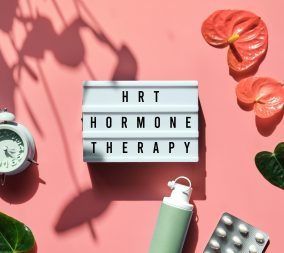 Text HRT Replacement Therapy on light box. Menopause, hormone therapy concept. Oestrogen replacement therapy awareness. Pink background with alarm clock, exotic leaves, pills, estrogene gel.