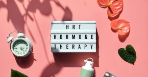 Text HRT Replacement Therapy on light box. Menopause, hormone therapy concept. Oestrogen replacement therapy awareness. Pink background with alarm clock, exotic leaves, pills, estrogene gel.